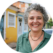 Retrato de mulher sorrindo, cabelos grisalhos cacheados, blusa verde e colar, em frente a casas coloridas.